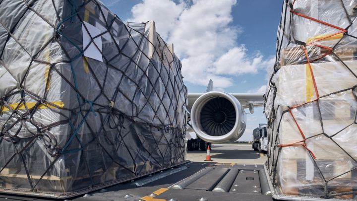 Cargo bundles secured for loading near an aircraft engine on a runway under a clear blue sky.