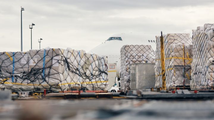 Pallets of cargo are stacked at an airport while a large airplane is seen in the background under cloudy skies.