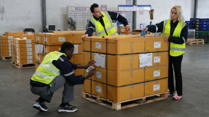 Three workers in reflective vests handle boxes in a warehouse, inspecting and organizing shipments.